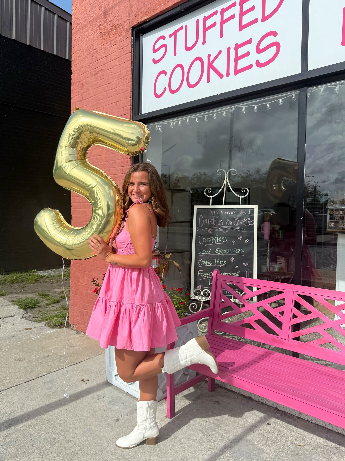 Woman in pink dress holding a gold number five balloon in front of a 'Stuffed Cookies' store.