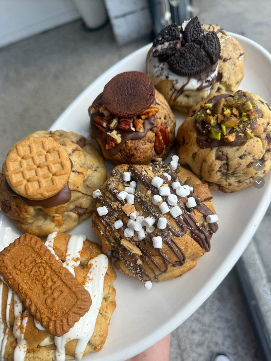 A white oval platter filled with assorted baked cookies and pastries, including chocolate chip cookies, sandwich cookies, and drizzled chocolate desserts, photographed from above.