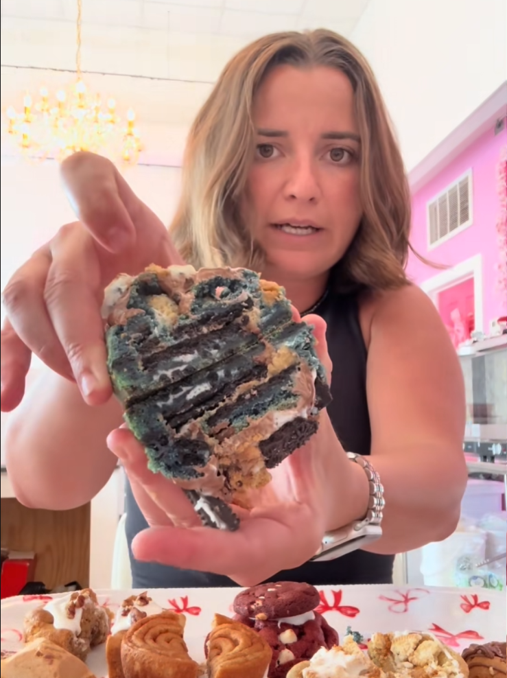 A woman holding up a thick, layered chocolate cake slice with blueberry jam in a bright kitchen setting.